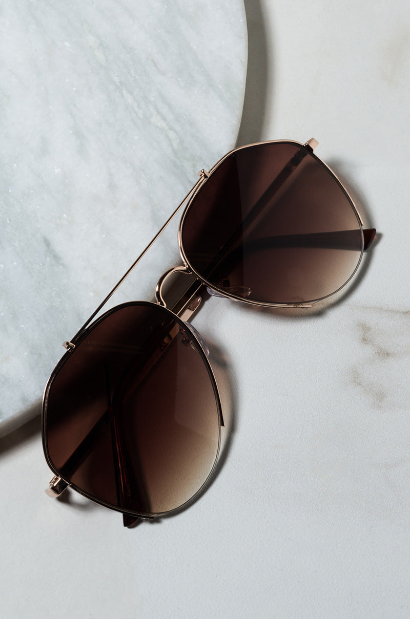 A pair of DEEP AVIATOR SUNGLASSES with gold frames and brown-tinted lenses is placed on a light marble surface. The sunglasses feature a double bridge and sleek design, subtly reflecting light. The background showcases textured circular marble decor.