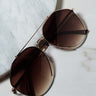 A pair of DEEP AVIATOR SUNGLASSES with gold frames and brown-tinted lenses is placed on a light marble surface. The sunglasses feature a double bridge and sleek design, subtly reflecting light. The background showcases textured circular marble decor.