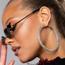 A person with slicked-back hair wears large New York Imports HOOP SILVER earrings and stylish dark butterfly-shaped sunglasses. They are dressed in a black top with red trim and hold their hand close to their face against a neutral background.