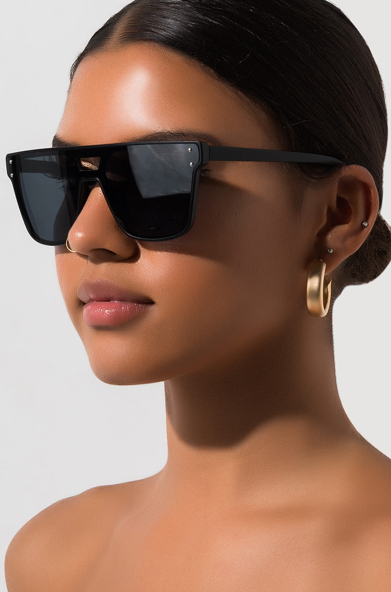 A person with dark hair pulled back, wearing large DOMO EVERYDAY SUNGLASSES and gold hoop earrings, is shown from the shoulders up against a plain background, looking slightly to the side.