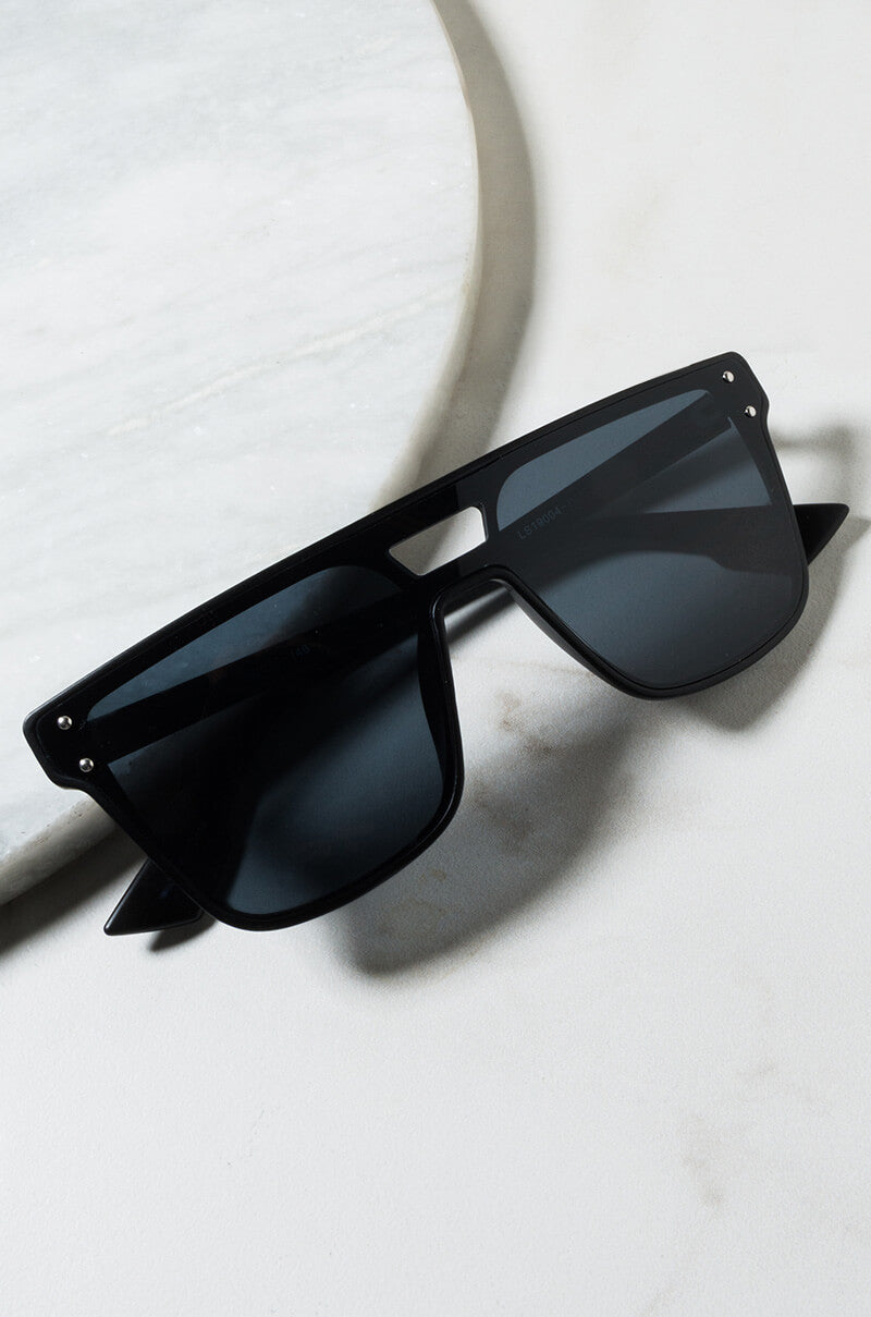 The DOMO EVERYDAY SUNGLASSES with rectangular lenses are placed on a light-colored marble surface. These sunglasses feature a minimalist design, sporting a solid black frame and dark tinted lenses.