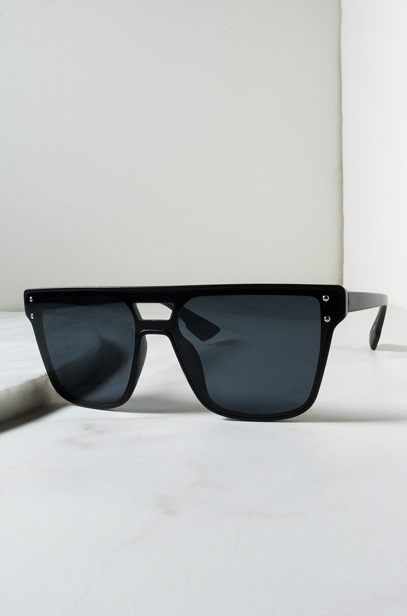 The DOMO EVERYDAY SUNGLASSES, a pair of black, oversized square sunglasses with dark lenses, are displayed on a light surface against a plain background. The frame is chunky with metal rivet detailing near the hinges, embodying a contemporary and minimalist aesthetic.