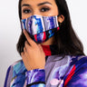 A person with long dark hair wears the DON'T @ ME FASHION FACE COVER, which features vibrant abstract imagery. They are wearing a matching top adorned with similar abstract patterns, and their hand rests on the face cover, partly masking their face. The background is white.