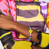 A person is wearing a vibrant, long-sleeved top featuring sections of bright pink, yellow, red, and black patterns. On their wrist is the "DONT FORGET TO SMILE BEADED BRACELET," adorned with a cheerful yellow smiley face charm. Their arms are crossed over their chest, revealing only the lower part of their face.