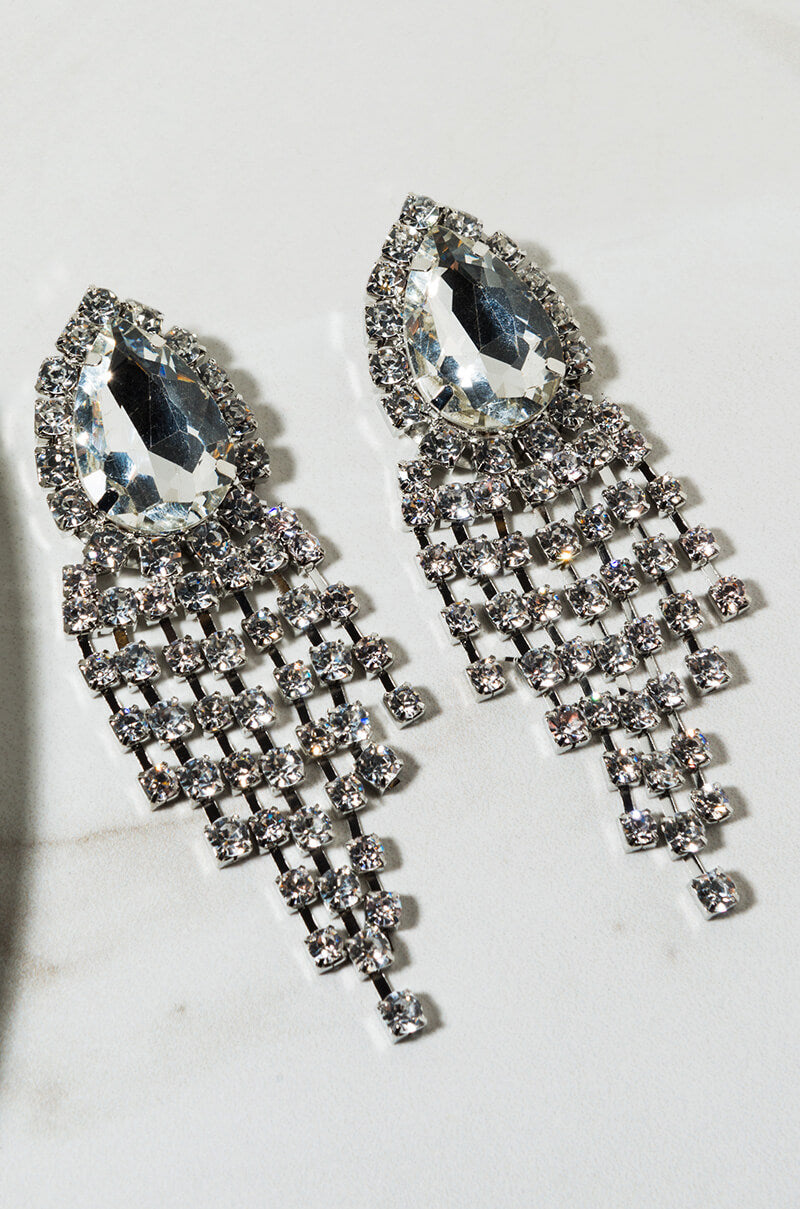 Introducing the EQUA DRIP EARRING: a pair of glamorous chandelier earrings adorned with large teardrop-shaped crystals, encircled by shimmering smaller round crystals. These earrings boast multiple layers of delicately dangling small crystals, creating a lavish and sparkling appearance that dazzles from any angle. The stunning design is beautifully showcased on a light surface, highlighting their exquisite craftsmanship.
