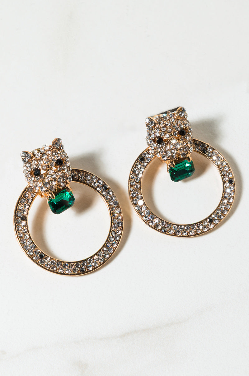 A pair of gold-toned EARRINGs designed as leopard heads encrusted with small, clear rhinestones. Each leopard head holds a larger green gemstone and both are attached to textured gold hoops adorned with more clear rhinestones. They are set on a light surface.