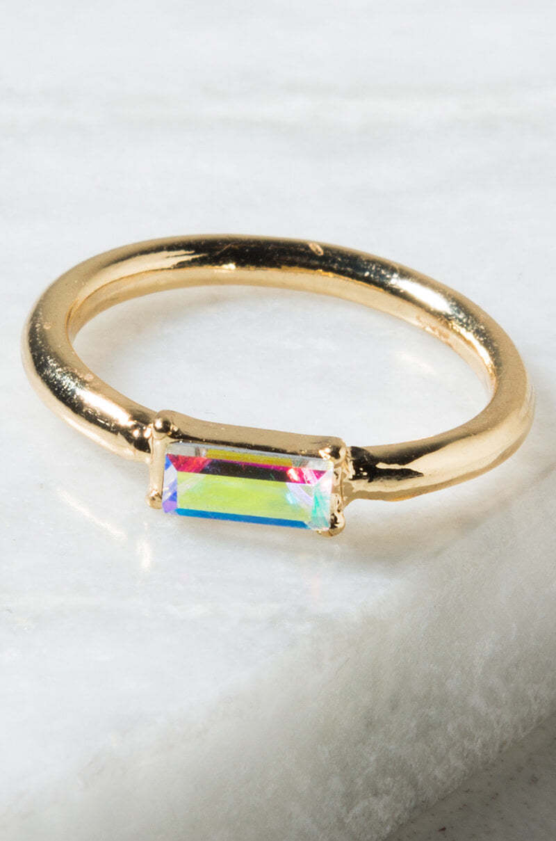 The IF I FELL AB RING, showcasing a rectangular, prism-like gemstone with a captivating rainbow spectrum of colors, is elegantly presented on a white marble surface. This gold ring features a simple and elegant band with the gemstone securely held in place by four prongs.