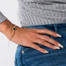 A person wearing a ribbed, long-sleeve gray shirt and fitted blue jeans rests their hand on their hip. Their nails are painted white with decorative patterns, and they are wearing the ROSE LINK BRACELET adorned with a dark green gemstone on their wrist.