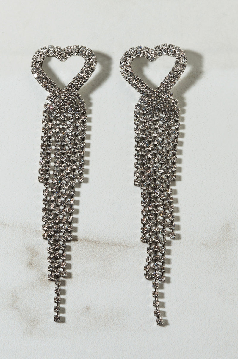 The HANDSY HEART EARRING features long, dangling earrings with heart-shaped tops adorned with small, shimmering rhinestones. Multiple strands of rhinestones cascade down from each heart, creating a sparkling and elegant design against a neutral background.