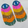 The EVIL SUMMER EYE FRINGE EARRING set features a pair of colorful handmade beaded earrings on a white surface, each showcasing an eye design at the top in blue and white, followed by rows of beads in pink, yellow, brown, orange, and ending with green and gold fringes.