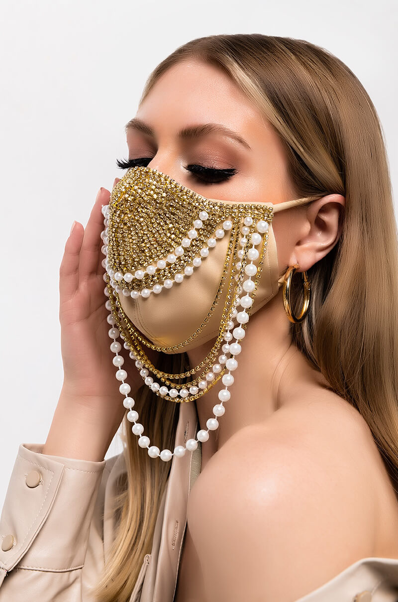 A woman with long, blonde hair is wearing the EXPENSIVE TASTE EXPENSIVE BITCH FACE MASK, which is beige and adorned with intricate jewel and pearl decorations, including hanging chains of pearls and gold beads. She is dressed in a beige jacket and gold hoop earrings, set against a plain white background.