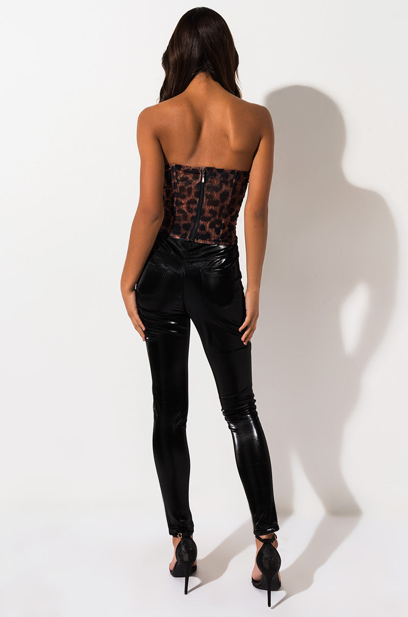 A person with long hair stands facing away from the camera, wearing a strapless leopard print top and SEXY LIQUID VELVET TROUSER PANT. They are also wearing black high-heeled shoes. The background is plain and light-colored, casting a shadow of the person on the wall.