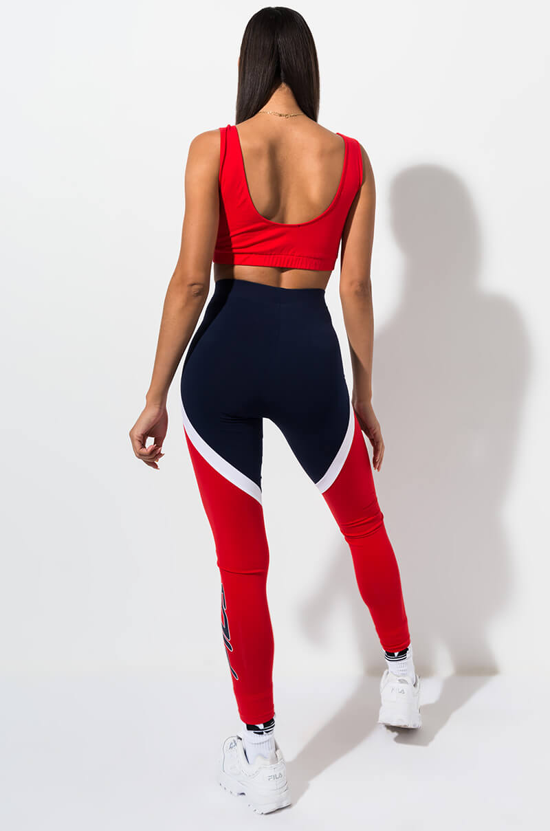 A person with long hair stands facing away, wearing a red sleeveless crop top and the FILA IVANNA HIGH WAIST LEGGINGS. The leggings are predominantly navy blue with red panels and white curved stripes along the sides. They are also sporting white sneakers.