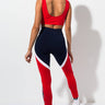 A person with long hair stands facing away, wearing a red sleeveless crop top and the FILA IVANNA HIGH WAIST LEGGINGS. The leggings are predominantly navy blue with red panels and white curved stripes along the sides. They are also sporting white sneakers.