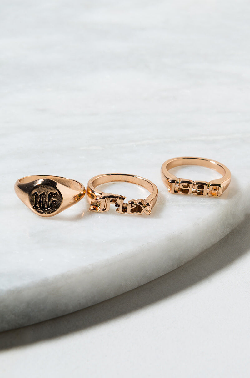 Three stunning rings from the FLEX 1990 RING SET displayed on a pristine white marble surface. The ring on the left showcases an intricately engraved seal, while the remaining two, placed side by side, boast bold geometric block designs.