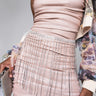 A person wearing the Florence Rhinestone Belt Skirt in shimmering pink, embellished with dangling rhinestones and featuring a ruched design, accessorized with a silver clutch. The wearer is also seen with a sheer, floral-patterned jacket.