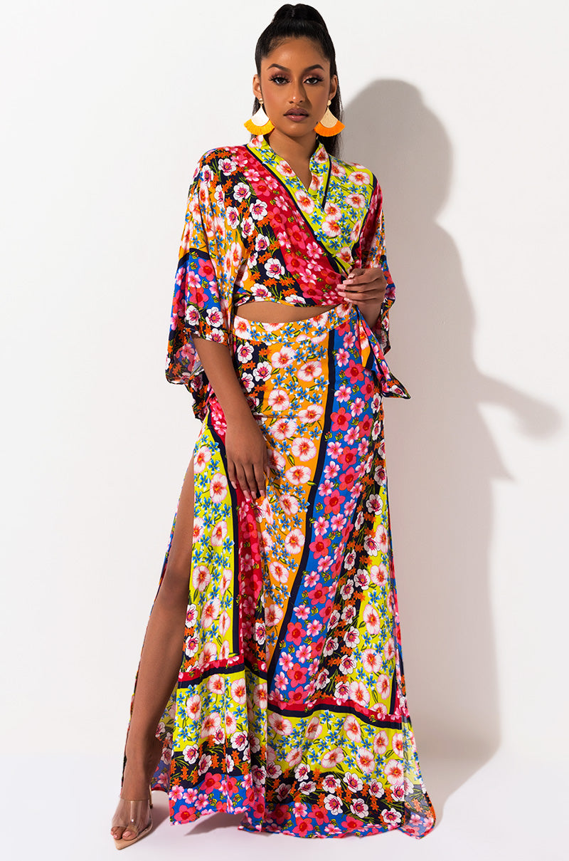 A woman stands against a white background, donning a vibrant SEXY FLORAL MAXI SKIRT with a slit on one side. She accessorizes with yellow statement earrings and clear high-heeled shoes. Her hair is styled in a high ponytail.