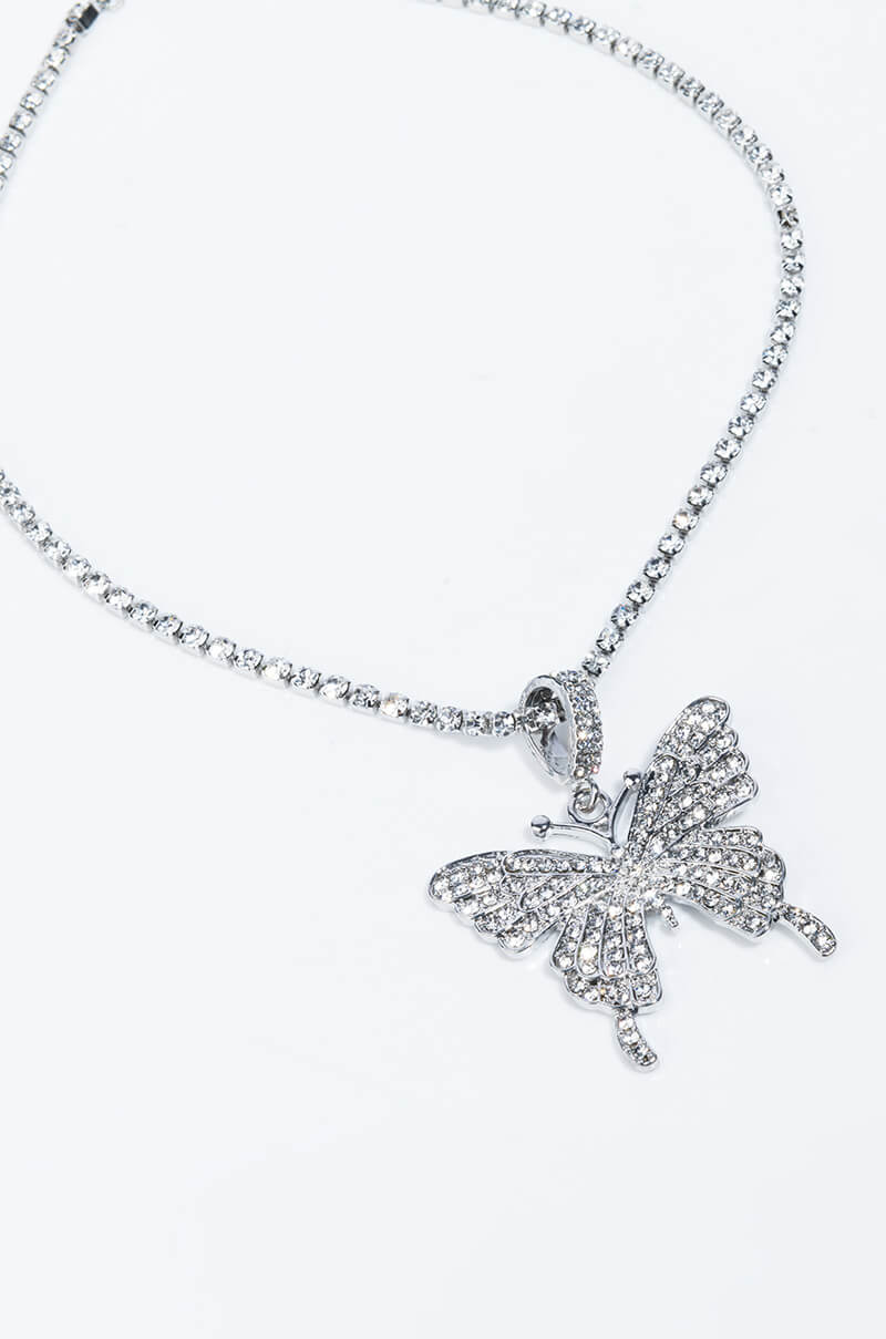 The FLY GIRL BLING BUTTERFLY NECKLACE showcases a butterfly-shaped pendant encrusted with sparkling gems. The pendant is elegantly suspended from a string of small, shimmering beads, giving it an elegant and delicate look. A plain, light-colored background accentuates the intricate details of the necklace.