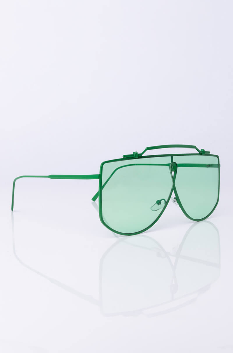 A pair of FLY HIGH SUNNIES with a thin green metal frame, double bridge, and green-tinted lenses rests on a reflective white surface. The stylish oversized sunglasses feature geometric lines and a modern aesthetic.