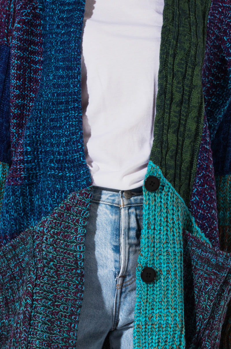 A person wearing the SEXY BOHO CARDIGAN LAYERING in vibrant shades of blue, green, and purple over a white T-shirt and light blue jeans. The cardigan features a mix of knit textures and two black buttons, partially covering the person's torso and hands.