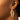 A close-up of a person showcasing the FRANCE HOOP EARRING. The hoop earring is gold-toned and features a textured rope design, standing out beautifully against the plain background. Only part of the person's face and ear is visible, drawing attention to the intricate details of the earring.