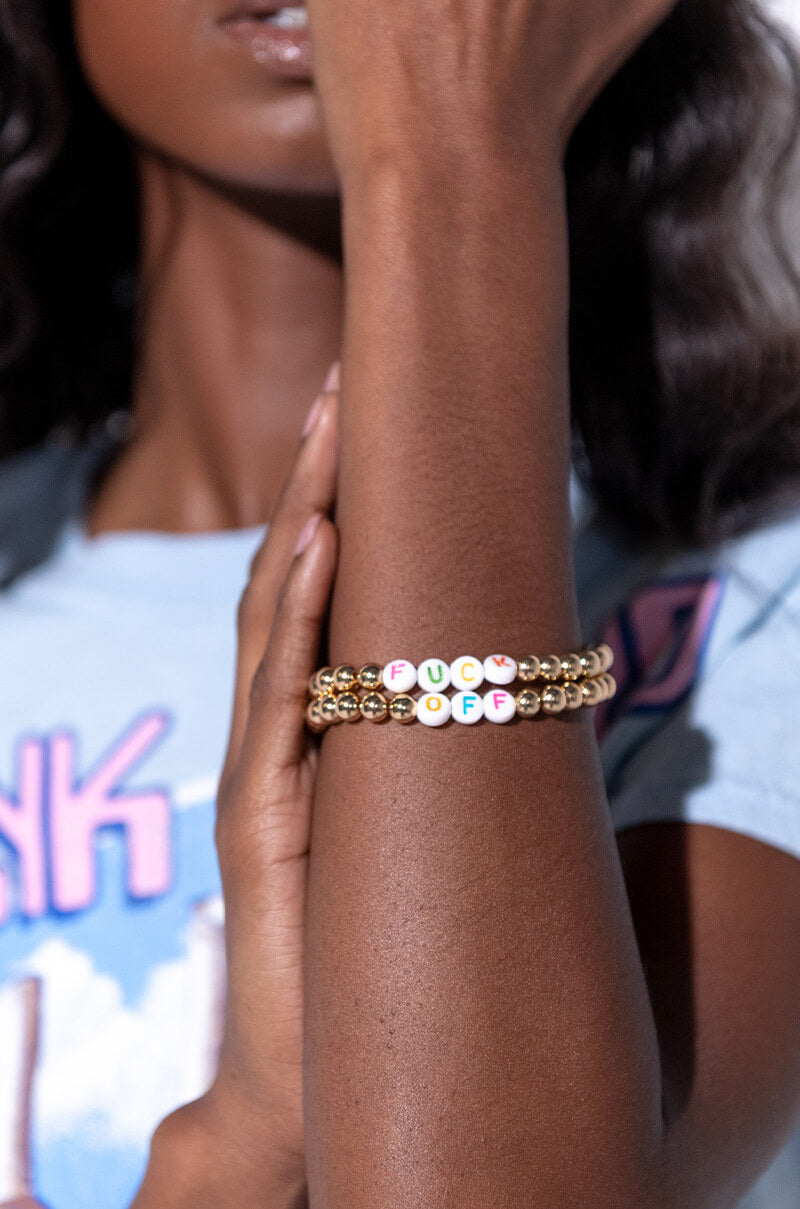 A person with a partially visible face is wearing a light blue shirt, holding an arm that displays the "FUCK OFF BEADED BRACELET" adorned with gold beads and colorful letter beads spelling out "FUCK OFF.