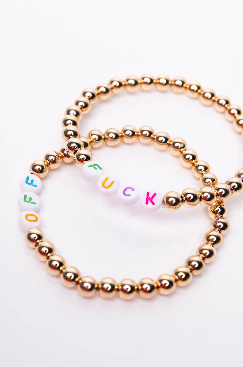 Two gold bead bracelets are intertwined. One bracelet includes white beads spelling "OFF" with a letter on each bead in different colors: green, yellow, and blue. The other bracelet includes white beads spelling "FUCK" in colorful letters, together forming the product name "FUCK OFF BEADED BRACELET.