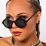 A person with braided hair wears large, round GAFAS TORTOISE SUNGLASSES and gold hoop earrings. They have a gold necklace featuring lion head pendants and a serious expression. The background is plain white.