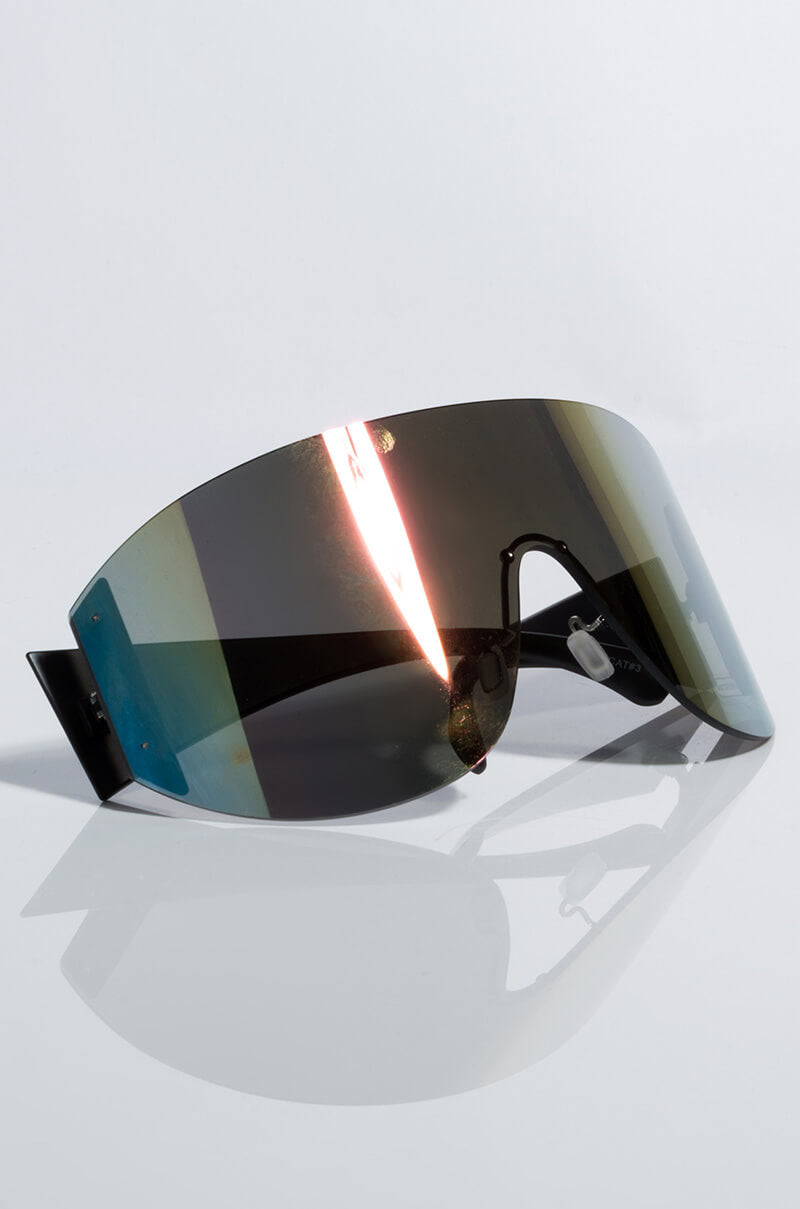 The JLO OMBRE SUNGLASSES feature a sleek, futuristic design with a single, large tinted lens extending across the front. The minimalistic frame includes black accents and an angular design. These sunglasses reflect light beautifully, resulting in a glossy appearance.