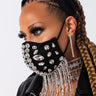 A woman wears a black face mask adorned with large rhinestones and draped chains. She also has a jeweled headband, long braided hair, dramatic eye makeup, and large dangling EARRING GOLD rhinestone earrings. She is dressed in a black jacket. The background is plain white.
