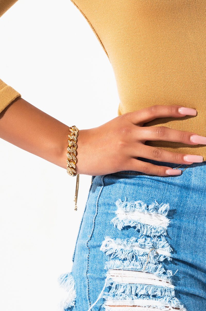 A person wearing a mustard-colored long-sleeve shirt and ripped blue denim jeans. Their left hand, adorned with the BRACELET gold chain bracelet, is resting on their hip. The background is white and uncluttered.