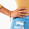 A person wearing a mustard-colored long-sleeve shirt and ripped blue denim jeans. Their left hand, adorned with the BRACELET gold chain bracelet, is resting on their hip. The background is white and uncluttered.