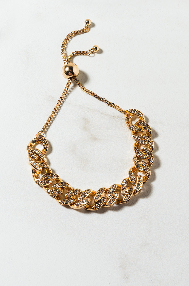 The BRACELET is a gold-toned chain bracelet with large links adorned with sparkling stones. It features an adjustable slider clasp with two small bead ends and is showcased on a white surface.