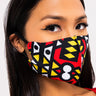 A woman with long dark hair is wearing the GOOD VIBES FASHION MASK COVER, which boasts a vibrant tribal pattern in red, yellow, black, and white geometric shapes. She has striking winged eyeliner and gazes directly into the camera with a neutral expression.