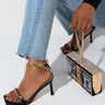 A person wearing light-wash, frayed-hem jeans and black high-heeled sandals with gold chain details is holding the GOT A STACK RHINESTONE MONEY CLUTCH in pink multi, designed to resemble a stack of dollar bills with golden and jeweled accents.