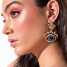 A woman with long brown hair wears the striking GRECIAN GLARE DANGLE EARRINGS, featuring a dark gemstone surrounded by smaller jewels. She has bold makeup with red eyeshadow and pink blush. She is facing to the side, with her ear and earring prominently displayed.