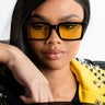 A person with long dark hair is wearing GROOVY BABY SUNNIES, which are large, rectangular sunglasses with yellow-tinted lenses, and a black and yellow jacket adorned with metal studs. They are leaning on their arm against a white background.