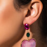 Close-up of a person wearing the HEART OF GOLD EARRING, which features a large pink gem at the top, a smaller green gem beneath it, and a heart-shaped pink pendant at the bottom. The person's ear and part of their face are visible against a plain background.