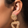 Close-up of a person's ear adorned with the HANA TEXTURE EARRING, featuring a golden, textured stud and a larger triangular, open pendant hanging below. The intricate and shimmering details are beautifully showcased against the person's smooth skin and pulled-back hair, highlighting the earring.