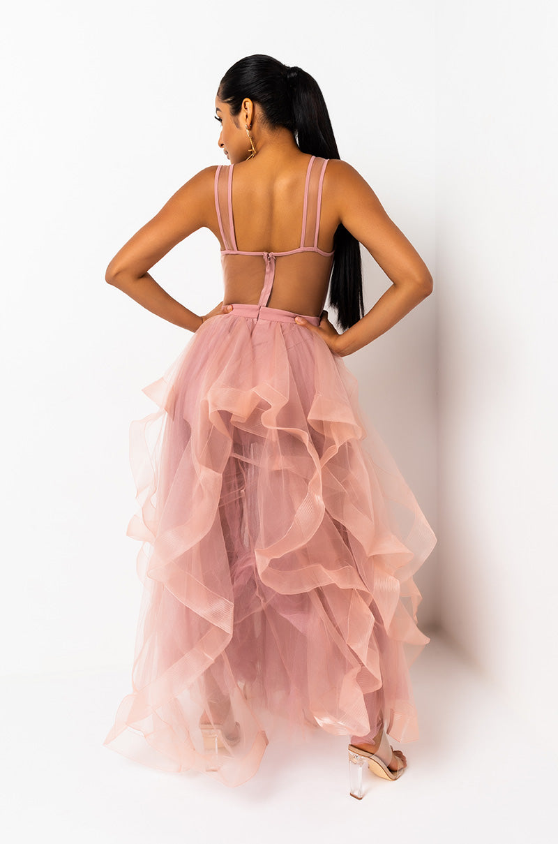 A woman stands with her back to the camera, wearing the HER WAY STRAPLESS MAXI DRESS, a pink, flowing, sheer dress with ruffled layers. Her hair is styled in a long ponytail as she poses in front of a white background while wearing clear high heels.