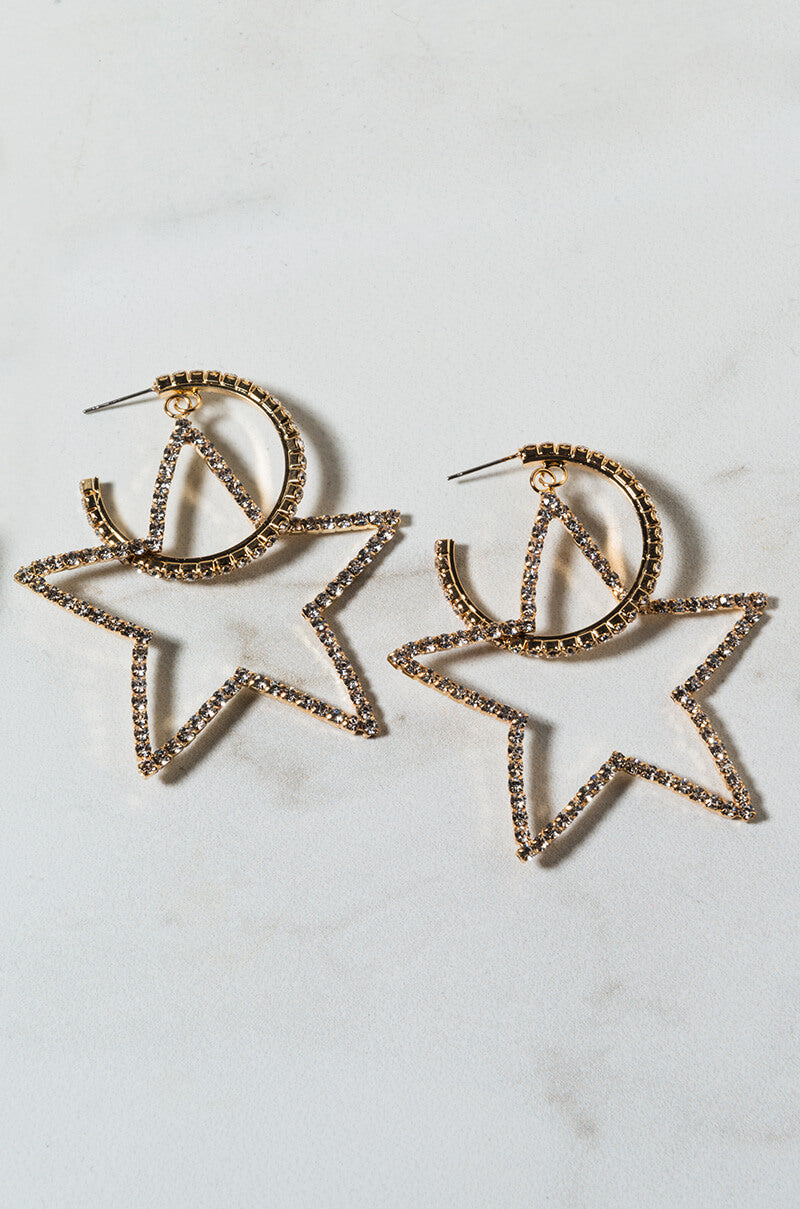 The SEXY HOOP EARRING is a pair of gold star-shaped hoop earrings adorned with small clear rhinestones. Featuring an open design, the circular loop at the top seamlessly transitions into the star shape. They are displayed on a light-colored, marble-like surface.