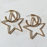 The SEXY HOOP EARRING is a pair of gold star-shaped hoop earrings adorned with small clear rhinestones. Featuring an open design, the circular loop at the top seamlessly transitions into the star shape. They are displayed on a light-colored, marble-like surface.