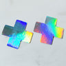 Two HOLOGRAPHIC X NIPPIE STICKER pasties in the shape of crosses, exhibiting a rainbow pattern. They are placed on a smooth white surface and display a reflective, iridescent sheen.