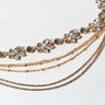 The HOMECOMING CHOKER features a delicate design with sparkling rhinestones and floral-shaped crystal clusters. Multiple gold chains of varying lengths drape elegantly from the main strand, creating a layered effect against a plain white background.