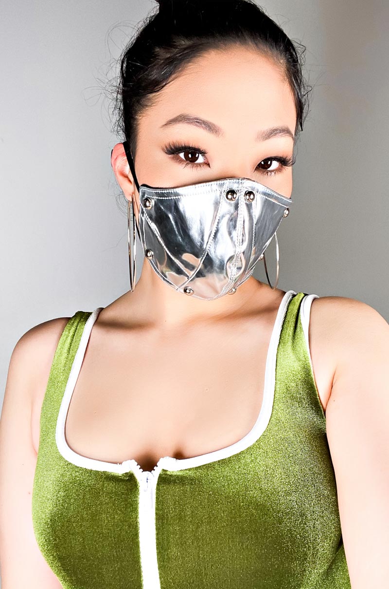 A person wearing a sleeveless, green velvet top with white trim and the HOT REFLECTIONS METALLIC PU FASHION FACE COVER. Large hoop earrings are visible, and the person's hair is tied up. The background is plain gray.