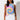 A woman with long, wavy black hair stands against a white background, wearing the I CANT EVEN TOP, a sleeveless tie-dye shirt featuring blue, yellow, and pink spiral patterns. She pairs it with high-waisted white jeans. Her expression is calm, and her arms rest naturally by her sides.