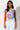 A woman with long, wavy black hair stands against a white background, wearing the I CANT EVEN TOP, a sleeveless tie-dye shirt featuring blue, yellow, and pink spiral patterns. She pairs it with high-waisted white jeans. Her expression is calm, and her arms rest naturally by her sides.