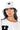 A person with long, dark hair wears the "I GOT SMILES BUCKET HAT," which is white and features a smiley face logo. They are dressed in a white shirt adorned with a black letter "H" patch, large hoop earrings, and have a serious expression on their face.