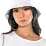 A person with long, dark hair wears the "I GOT SMILES BUCKET HAT," which is white and features a smiley face logo. They are dressed in a white shirt adorned with a black letter "H" patch, large hoop earrings, and have a serious expression on their face.