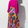 A woman stands with her back to the camera, wearing a vibrant, long-sleeved pink top and FASHION LEADER's colorful IN BLOOM PLEATED MAXI SKIRT featuring a multi-color floral pattern. She has blonde hair and is wearing clear high heels. The white background highlights her outfit.